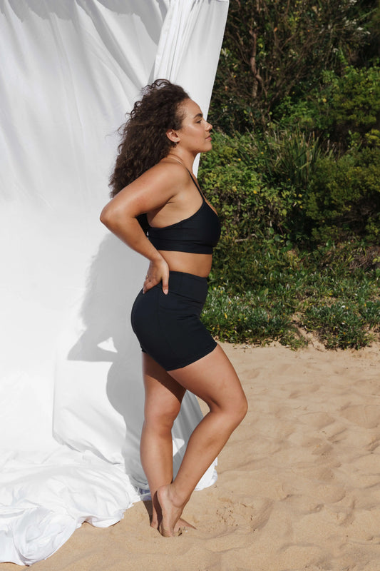 Model at the beach facing the side wearing our Inala Crop and Allawah Shorts in the colour Black